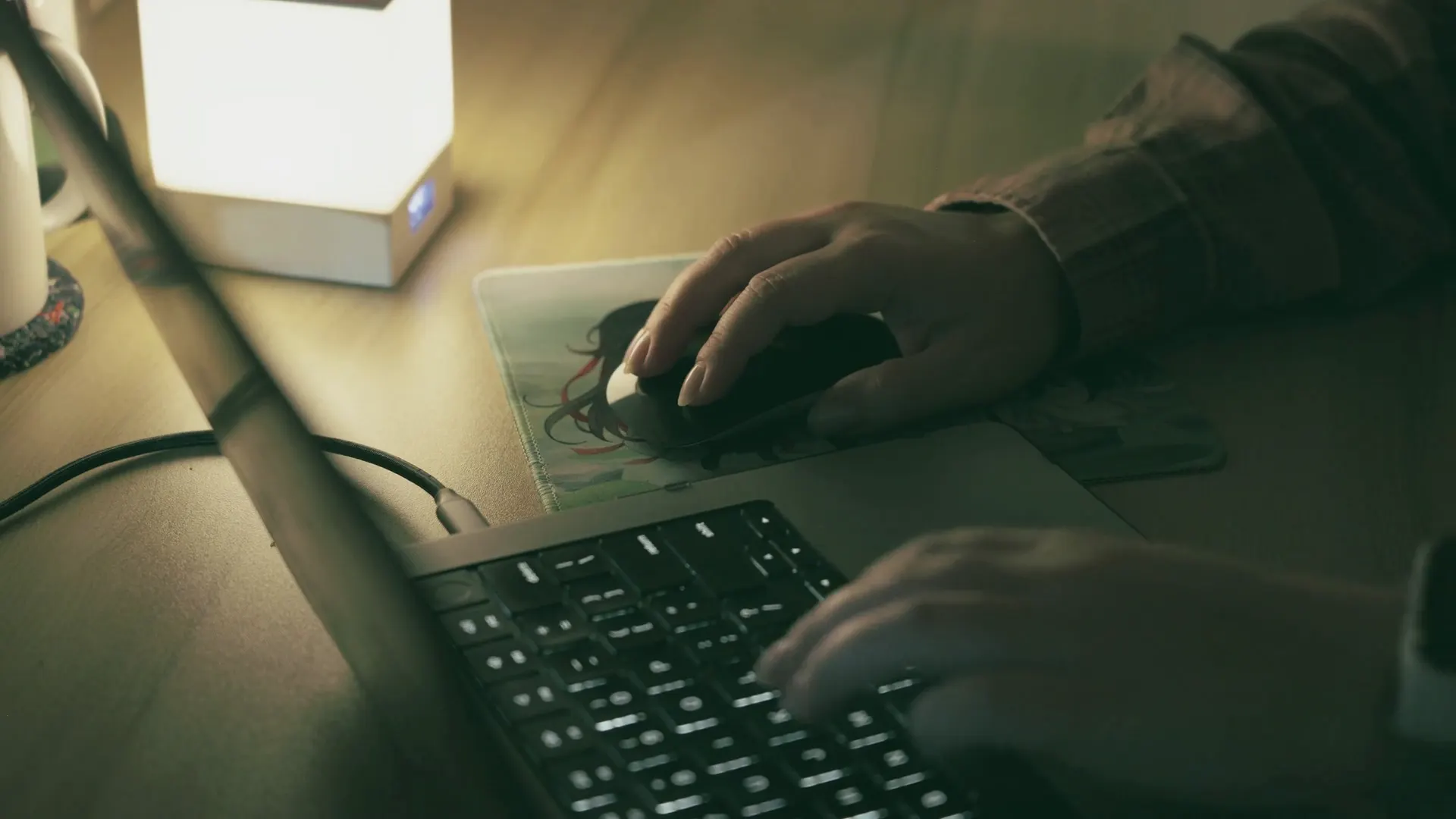 Close-up of hands on a laptop keyboard and mouse at a lamp-lit desk at night.